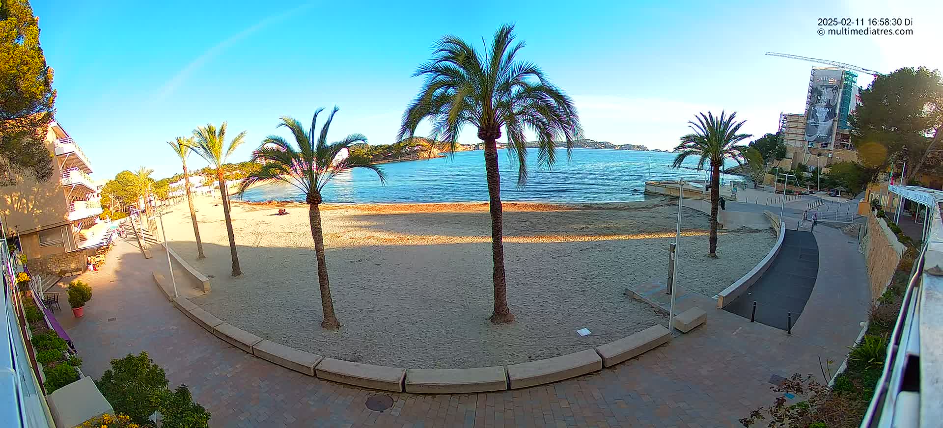 A sandy beach with palm trees and a paved walkway curves around it, overlooking calm ocean water under a clear, sunny sky.