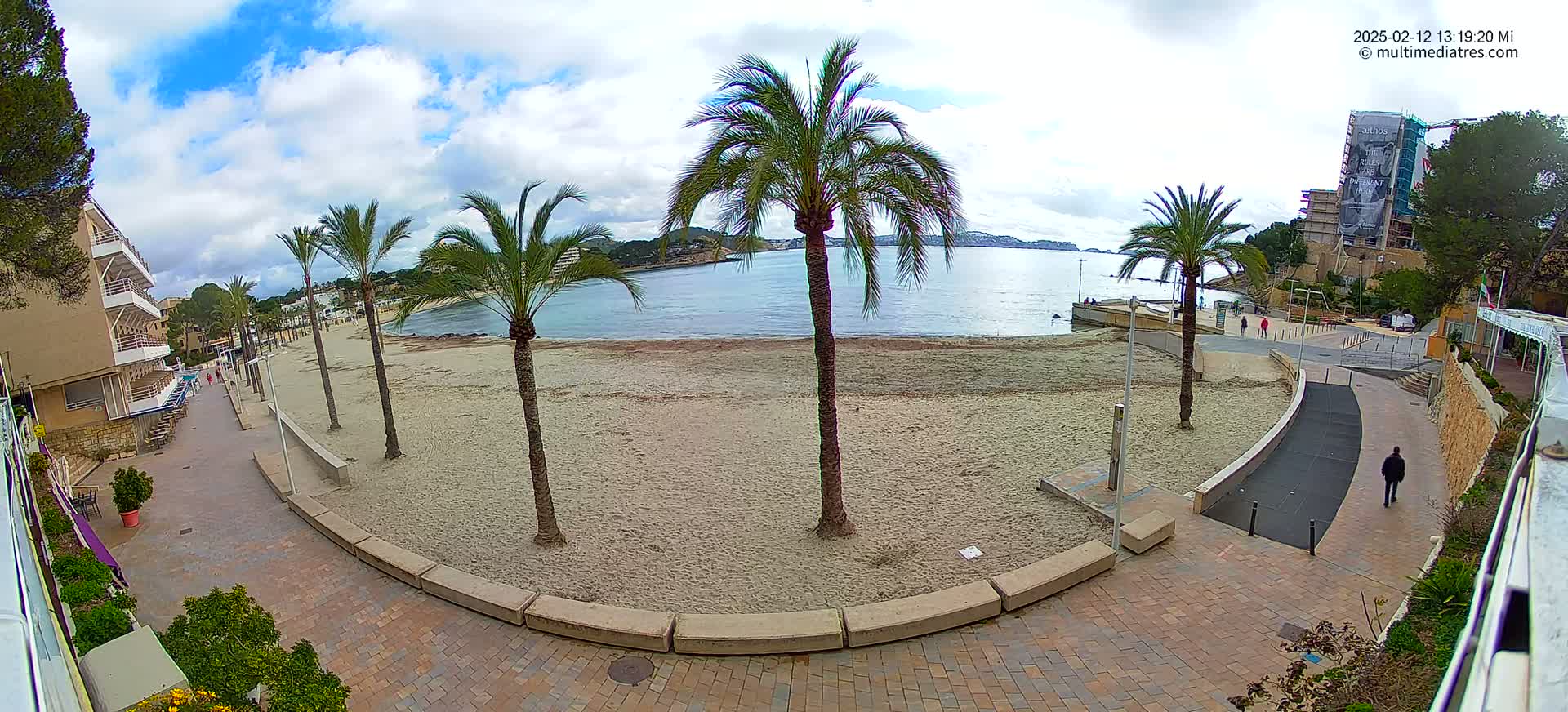 A mostly sunny day shows a sandy beach with palm trees, a paved walkway, and buildings along the coast.