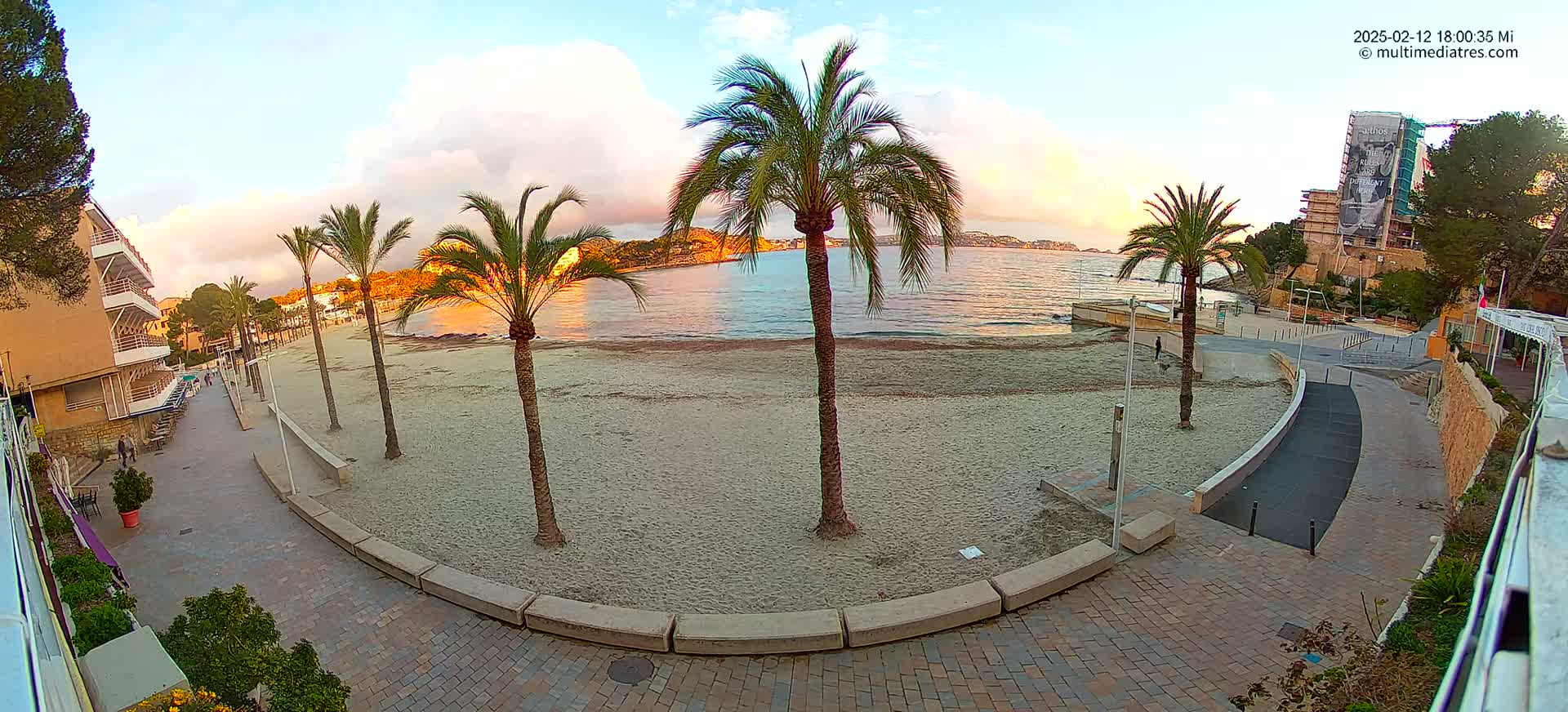 A sandy beach with palm trees is bordered by a paved walkway and buildings, under a partly cloudy sunrise sky.