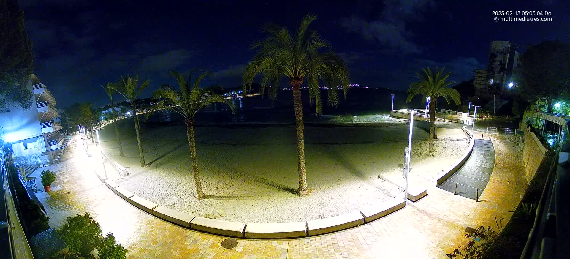 A nighttime view of a sandy beach at the edge of a paved walkway lined with palm trees and buildings under a partly cloudy sky.