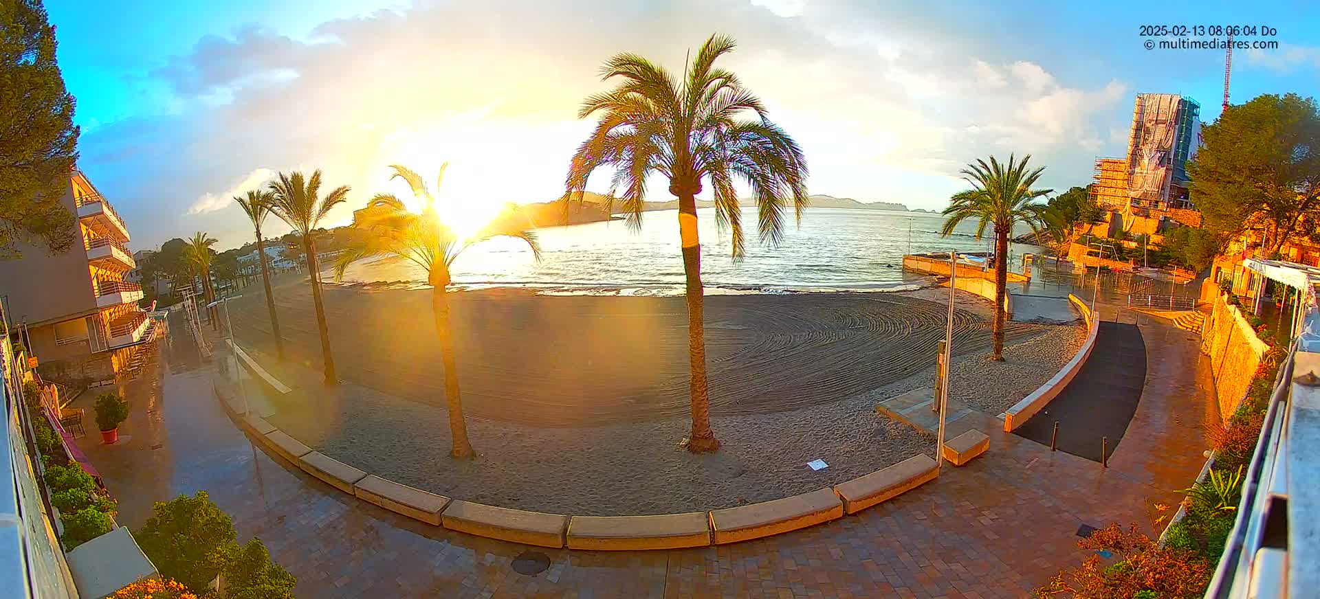 A sunrise illuminates a wet beach with palm trees, a building under construction, and a paved walkway.