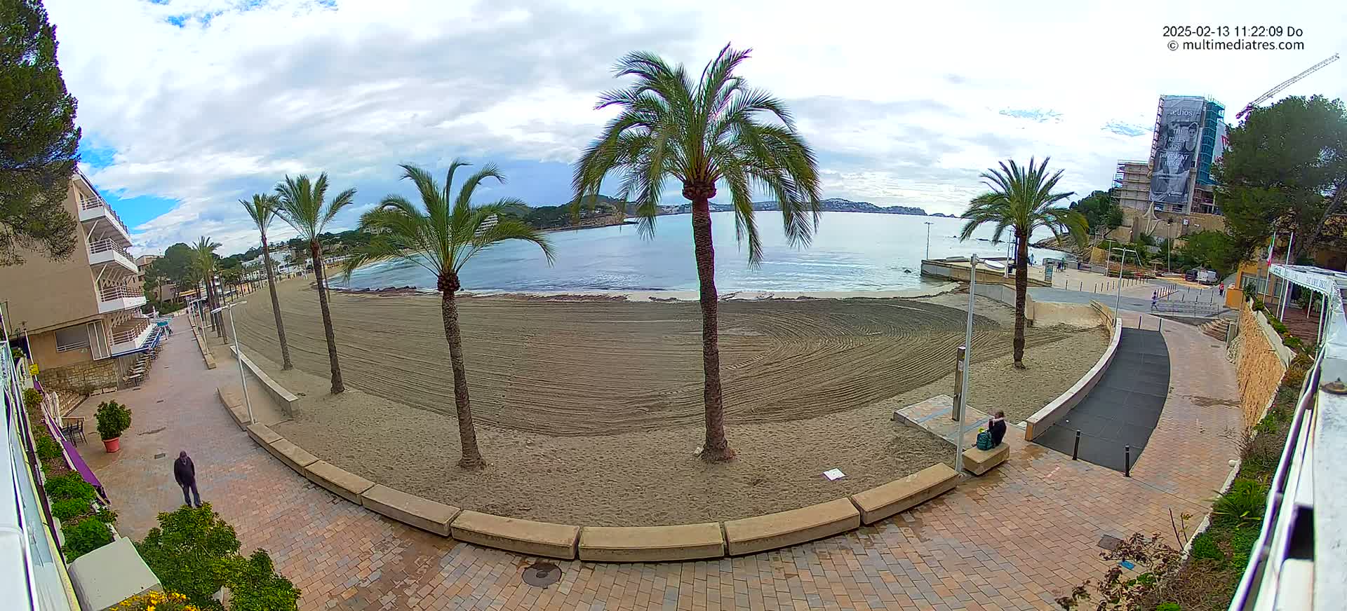 A mostly cloudy day reveals a sandy beach with palm trees lining a paved walkway, overlooking a calm bay with buildings and hills in the distance.