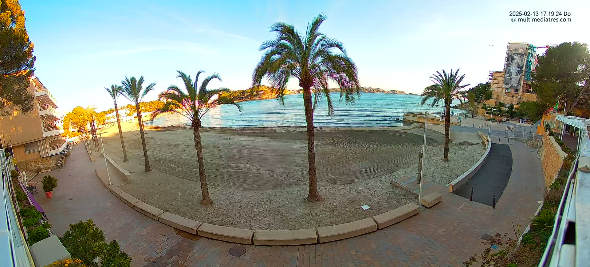 A sandy beach with palm trees and buildings is seen on a calm, sunny day.