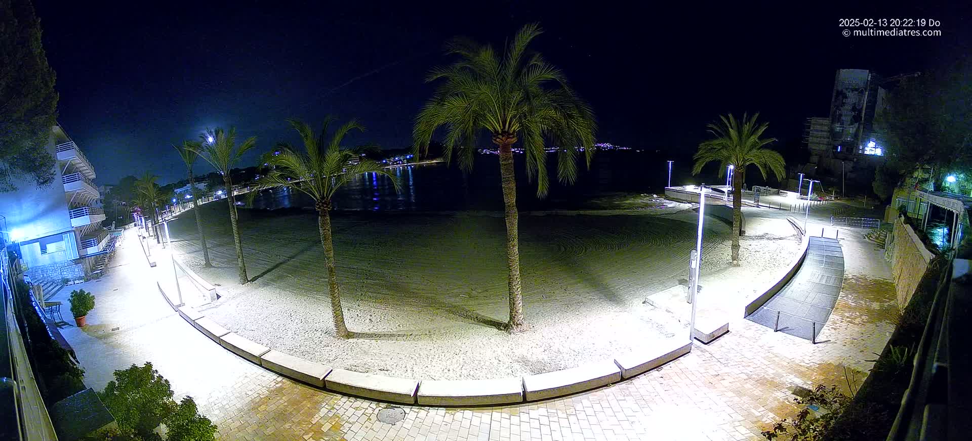 A nighttime view of a sandy beach at low tide, illuminated by artificial lights, with palm trees lining a paved walkway next to calm water.