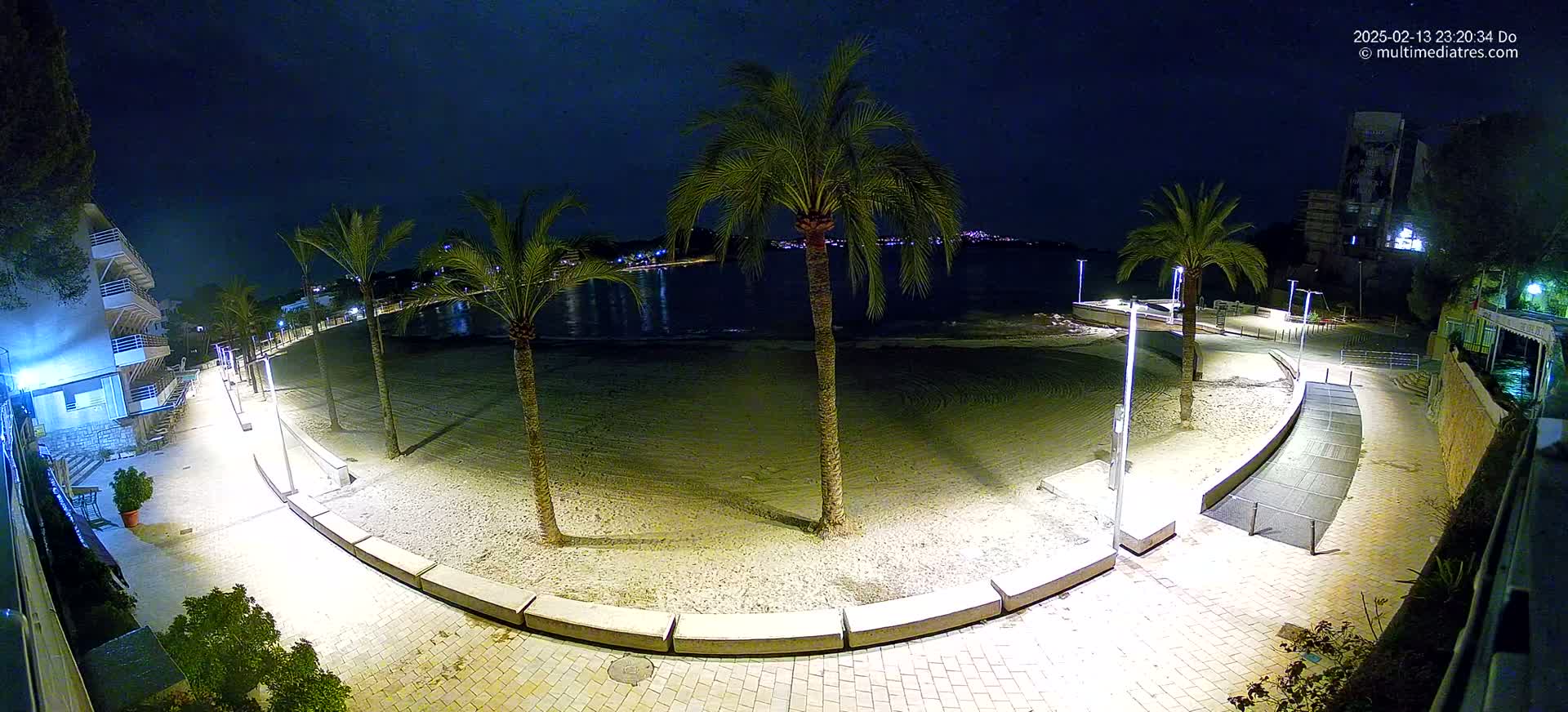 A nighttime view of a sandy beach with palm trees lining a paved walkway, illuminated by artificial lights, next to a calm body of water.