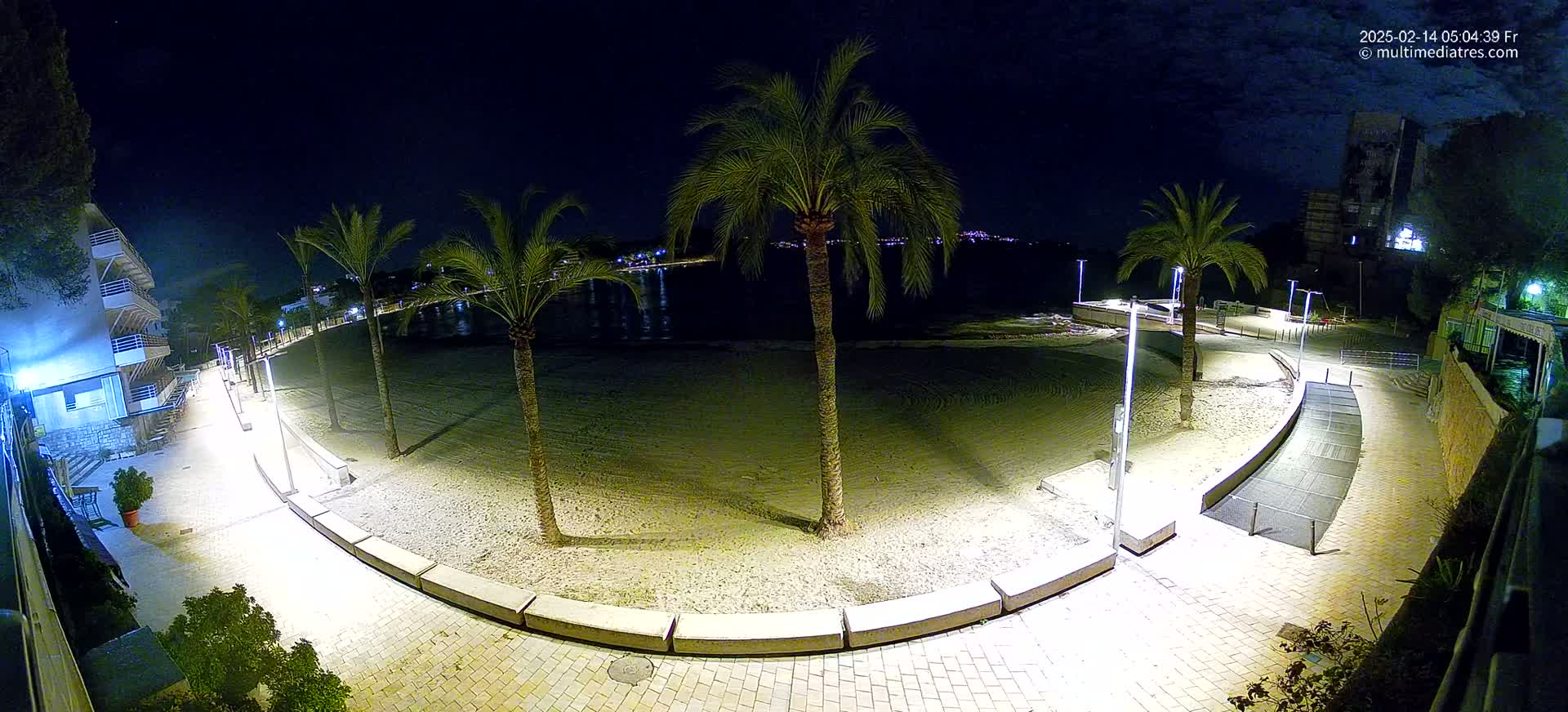 A nighttime, high-angle view shows a sandy beach bordered by a paved walkway with palm trees and lights, overlooking a calm body of water.