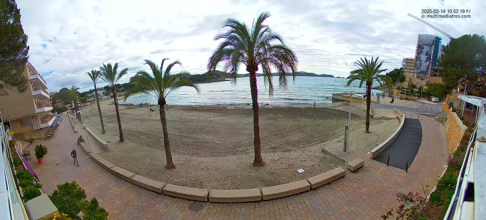A mostly sunny day shows a sandy beach with palm trees, a paved walkway, and buildings overlooking a calm sea.