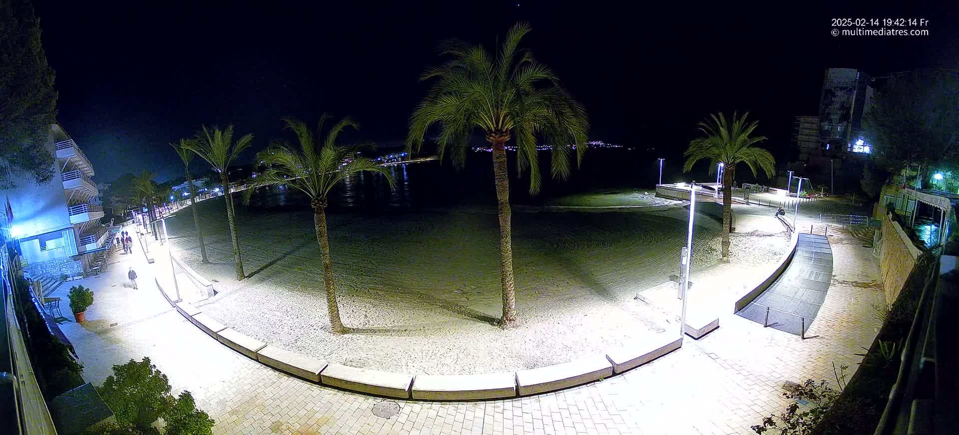 A nighttime, illuminated view of a sandy beach with palm trees, a paved walkway, and a few people walking along the shore.