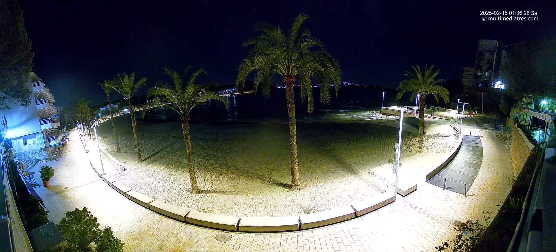 A nighttime view of a sandy beach with palm trees, illuminated walkways, and buildings along the shoreline under a clear, dark sky.
