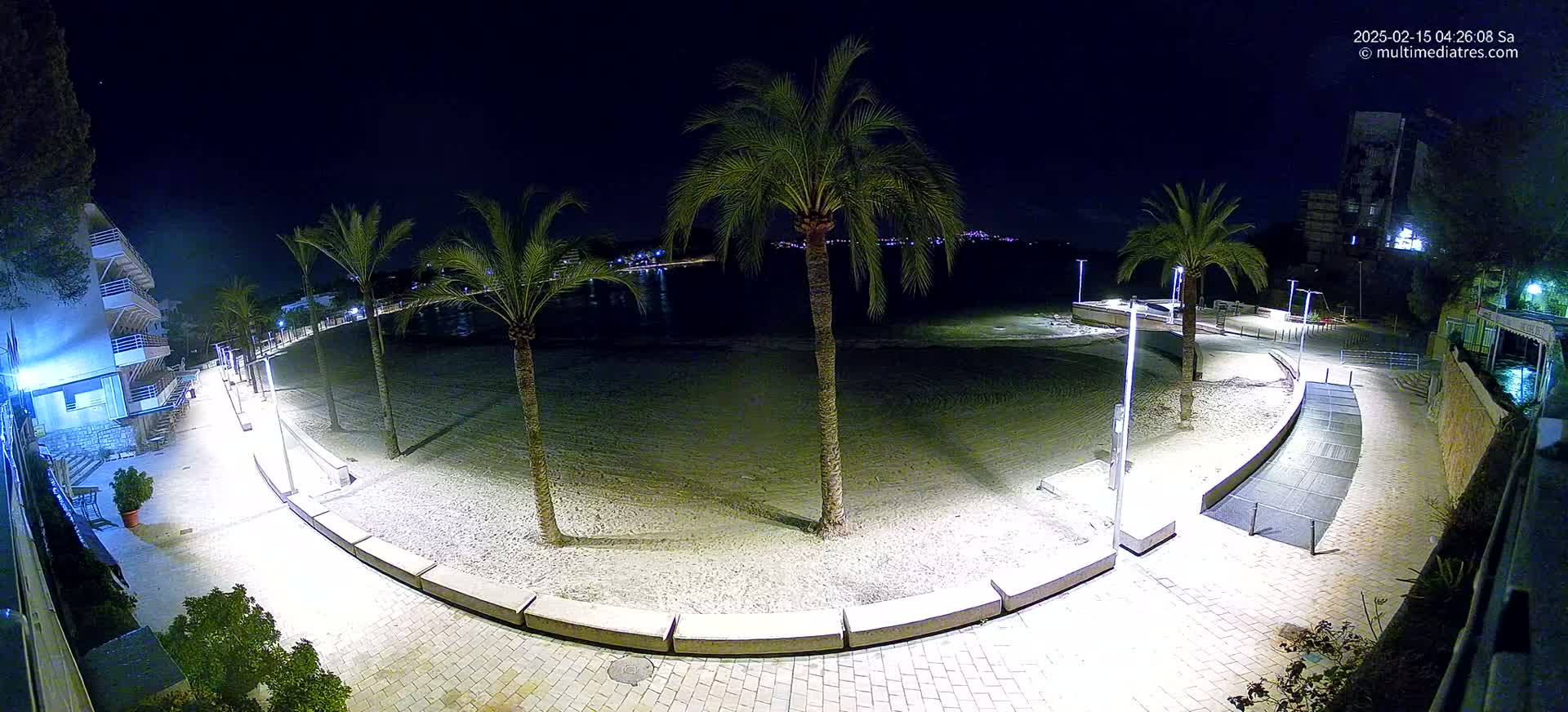A nighttime view of a sandy beach with palm trees and illuminated walkways, bordered by buildings.
