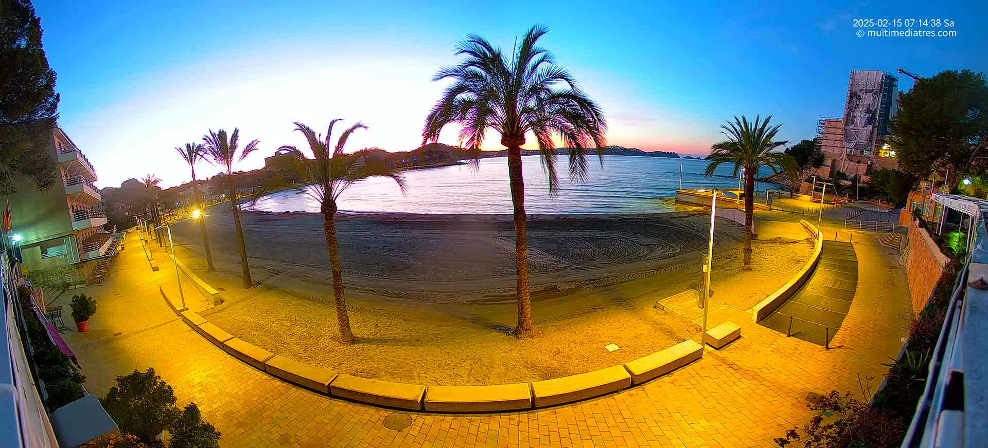 A sandy beach at sunrise is illuminated by artificial lights, with palm trees lining a paved walkway next to a calm sea.