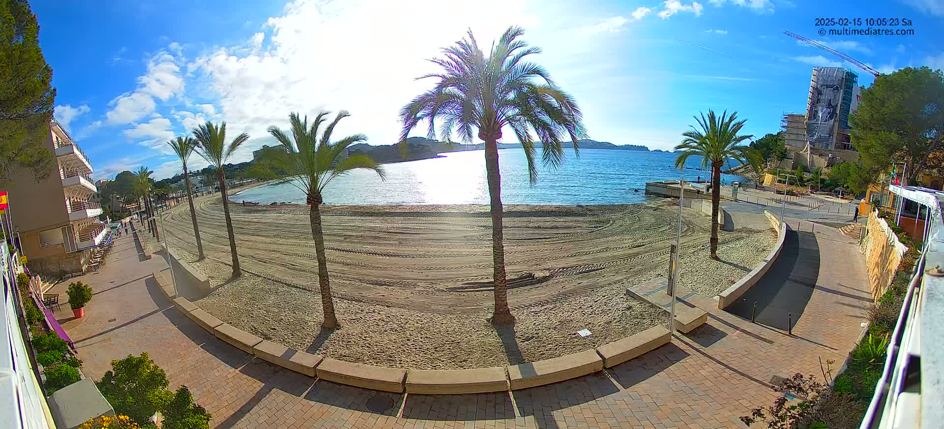 A sandy beach with palm trees and a paved walkway is visible on a sunny day, overlooking a calm body of water.