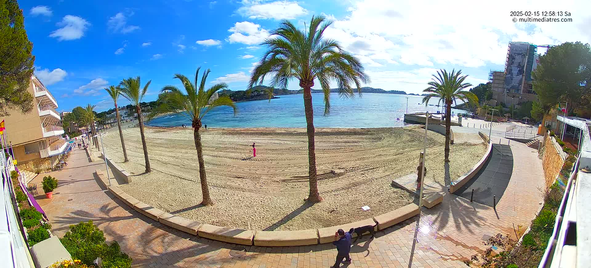 A sandy beach with palm trees and a paved walkway next to a calm, blue body of water under a mostly sunny sky.