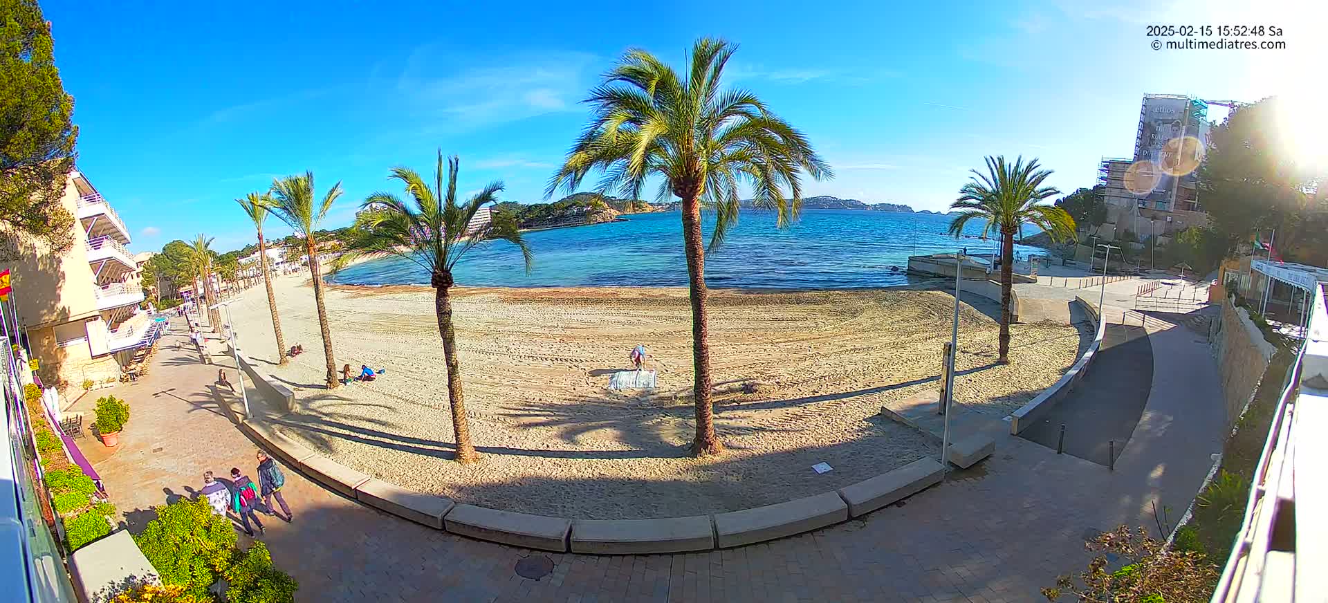 A sunny day at a sandy beach with palm trees, buildings, and a few people walking along a paved walkway.