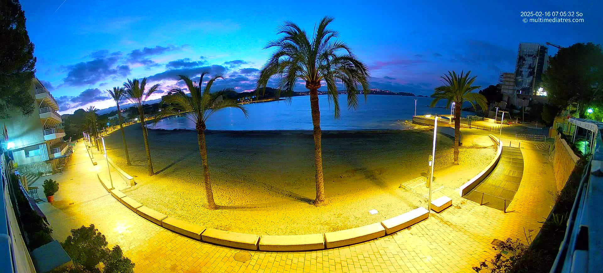 A nighttime view of a sandy beach with palm trees, illuminated walkways, and buildings along the shoreline under a twilight sky.