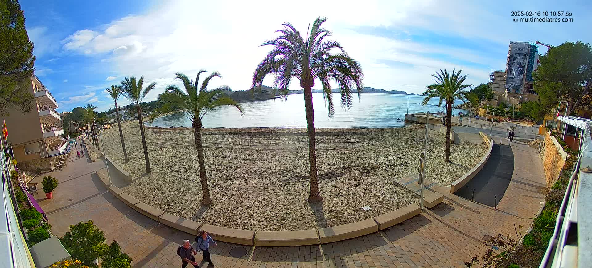 A sandy beach with palm trees lines a paved walkway next to a calm body of water under a mostly sunny sky.