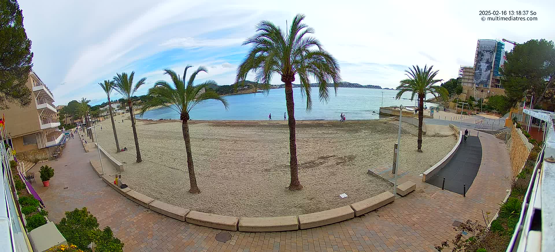A sandy beach with palm trees and a calm, blue bay is seen under a partly cloudy sky, next to a paved walkway and buildings.
