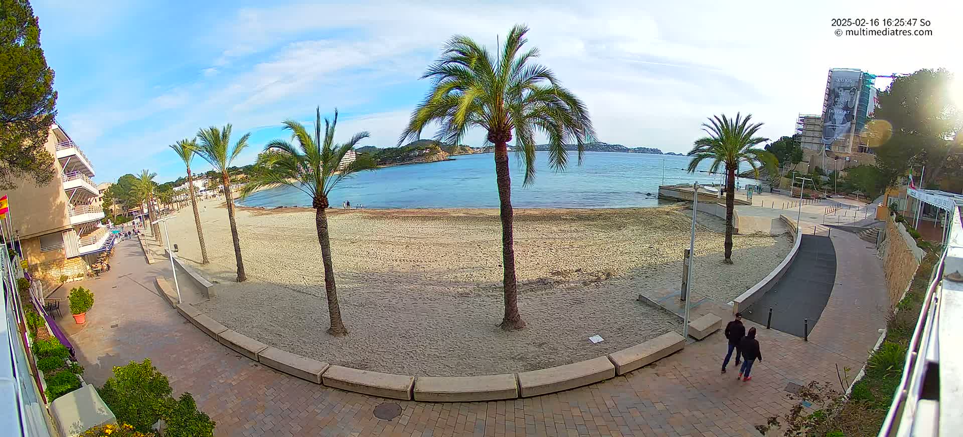A sandy beach with palm trees and a paved walkway next to a calm, blue ocean under a mostly sunny sky.