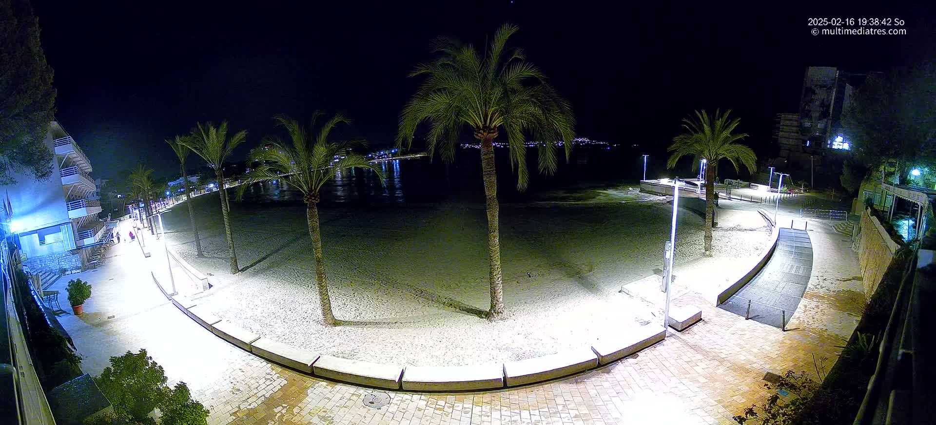 A sandy beach at night is lit by artificial lights, bordered by a paved walkway lined with palm trees and buildings.