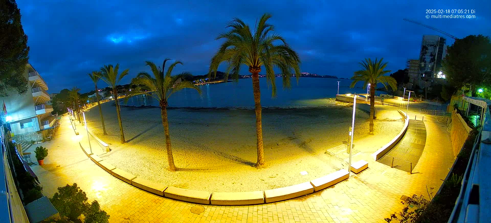 A nighttime view of a sandy beach with palm trees, illuminated walkways, and buildings along the waterfront under a dark blue, cloudy sky.