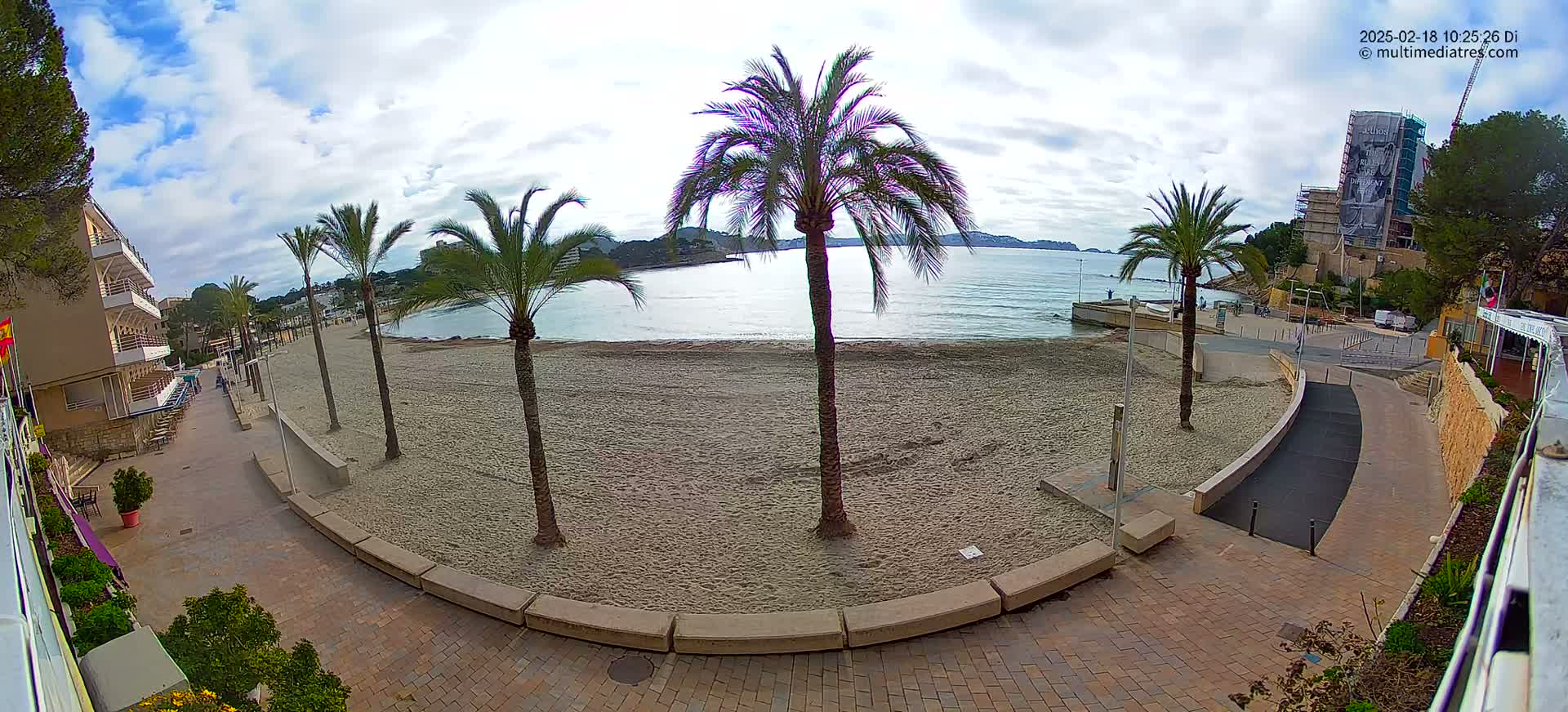 A mostly sunny day on a sandy beach with calm water, palm trees, and buildings in the background.