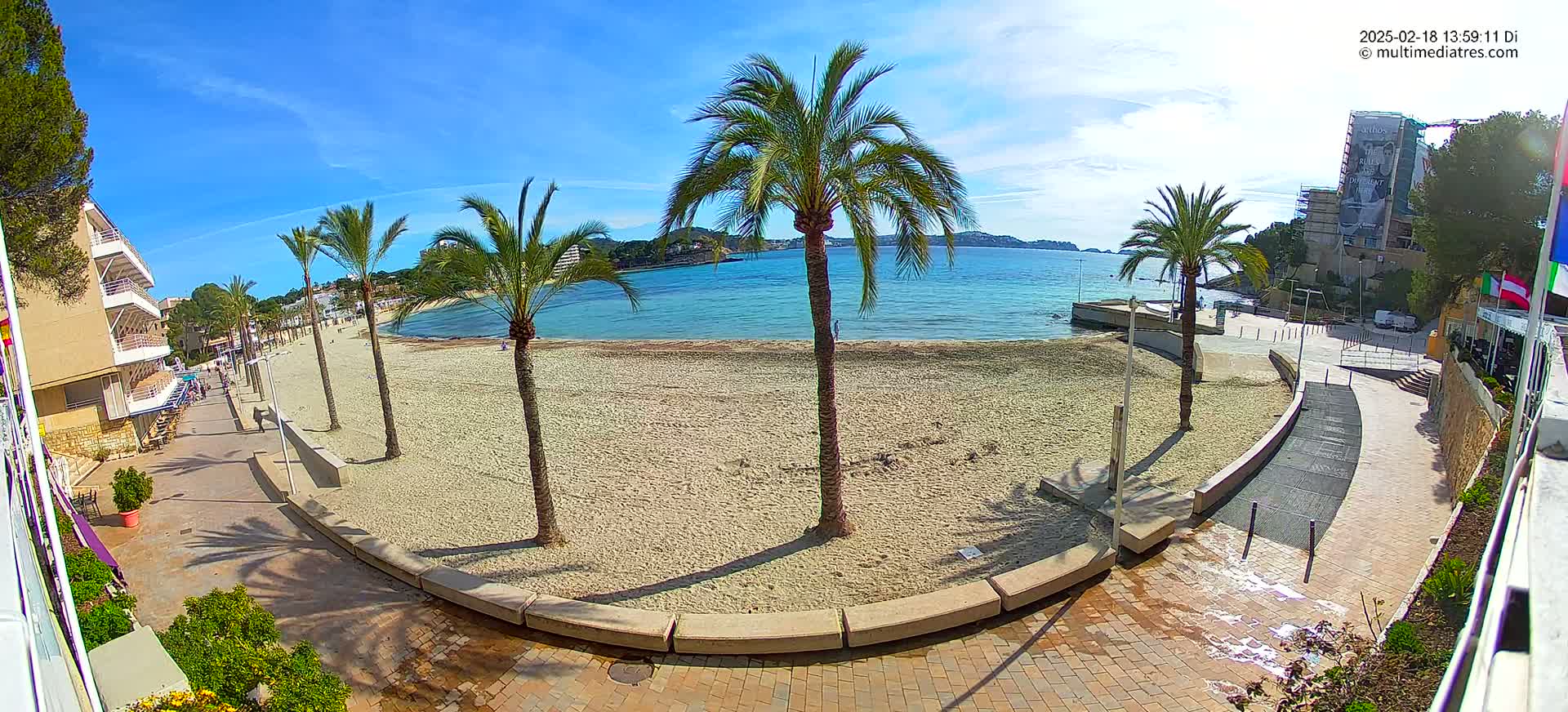 A sandy beach with palm trees lines a paved walkway next to calm, light-blue water under a sunny sky.