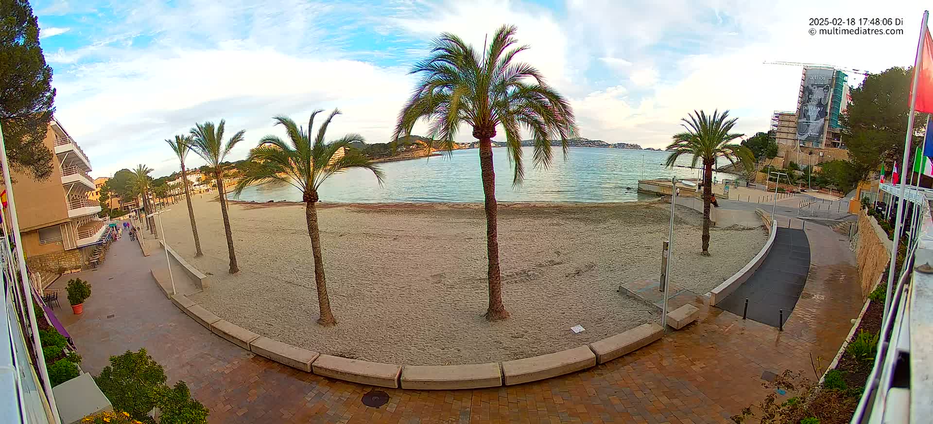 A sandy beach with palm trees lines a paved walkway next to a calm body of water under a partly cloudy sky.