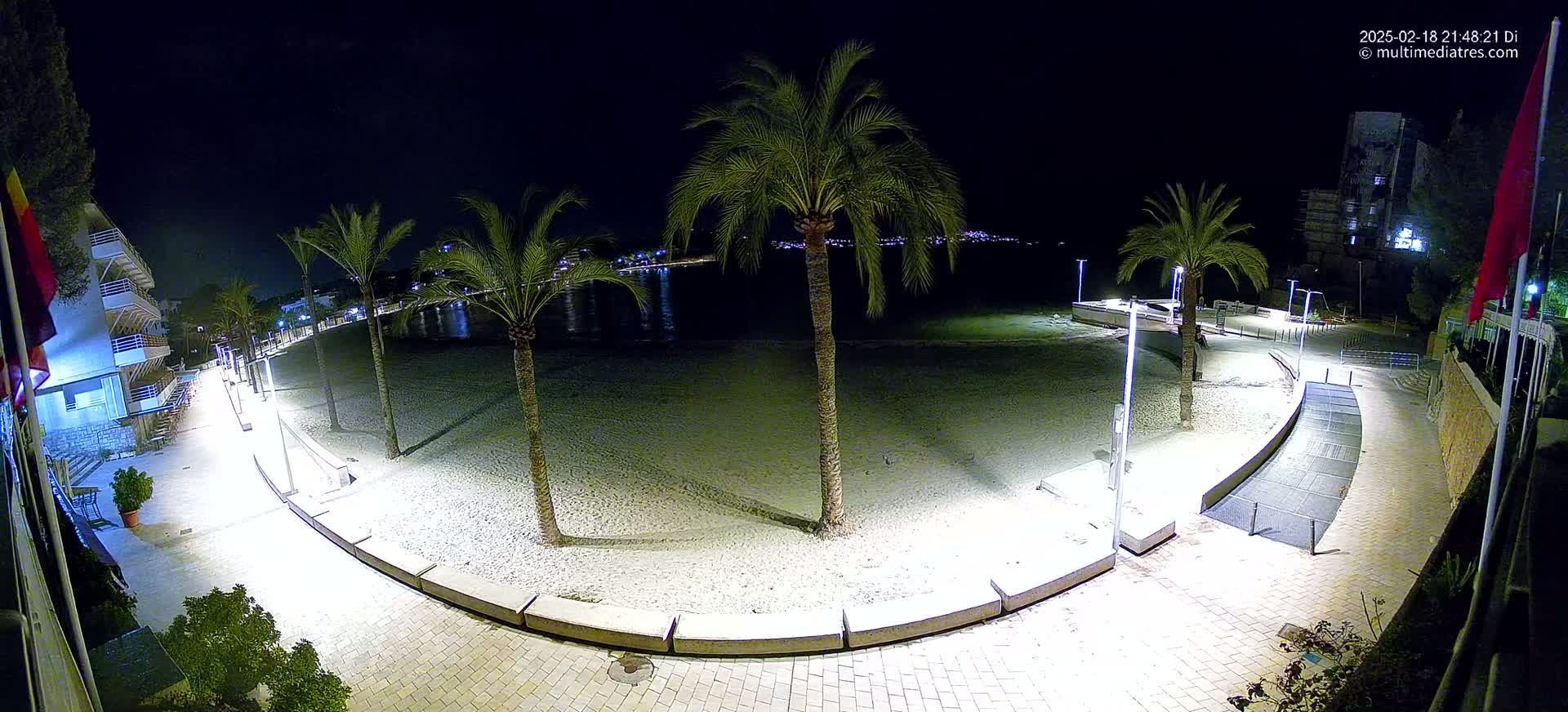 A nighttime, illuminated view of a sandy beach with palm trees, bordered by a paved walkway and buildings.