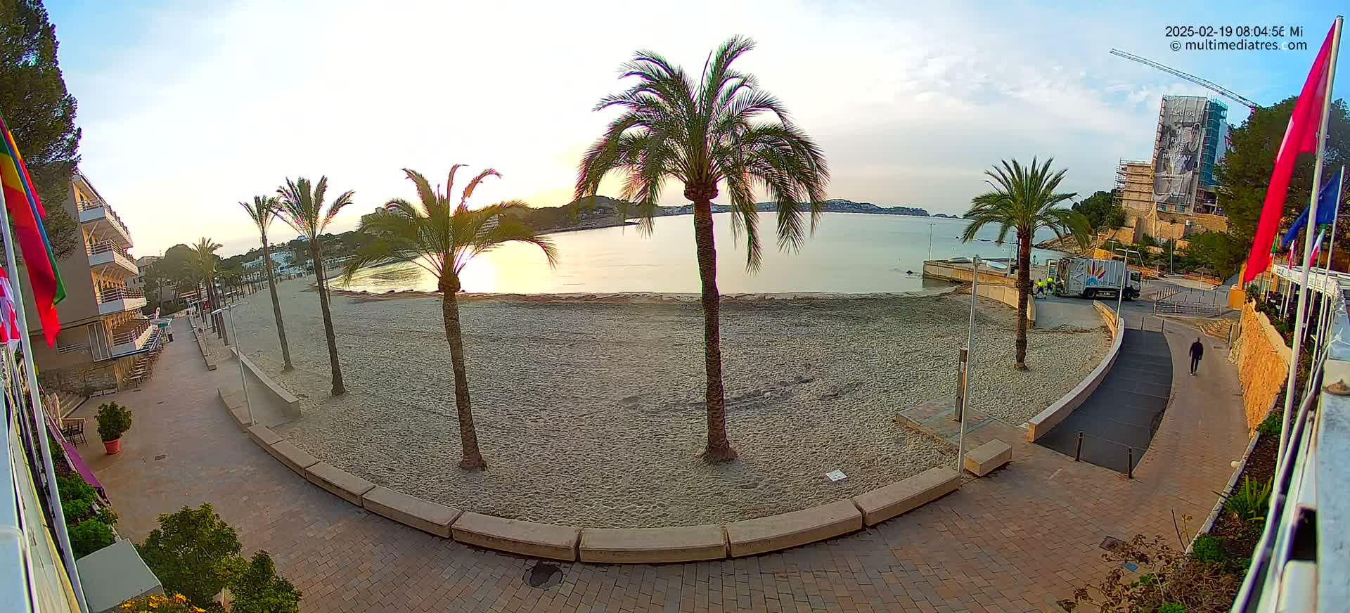 A sandy beach at sunrise, bordered by palm trees and a paved walkway leading to a building, with calm water in the background.