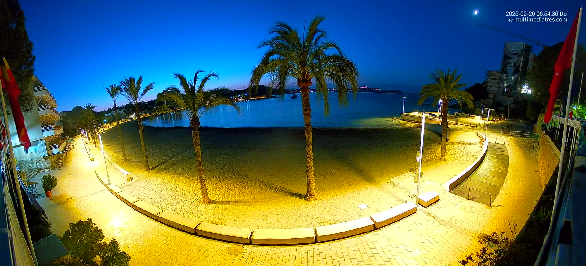 A brightly lit, curved walkway at night next to a sandy beach bordered by palm trees and calm water under a twilight sky.