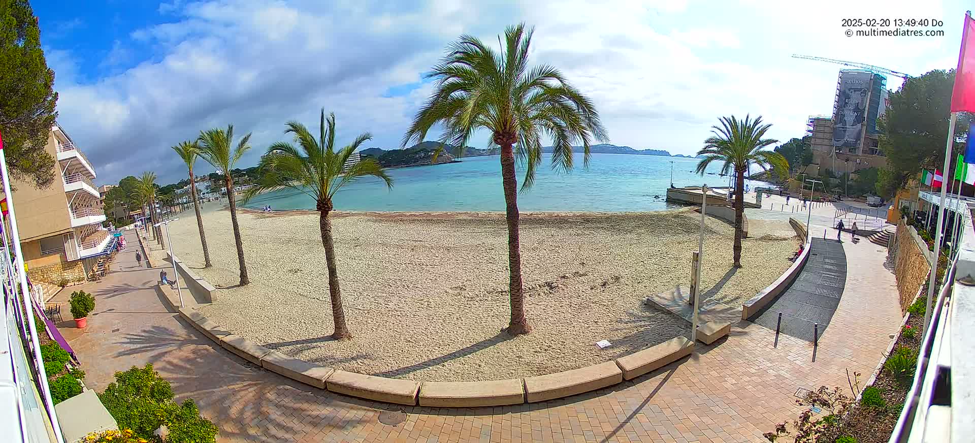 A mostly sunny day reveals a sandy beach bordered by palm trees, a paved walkway, and buildings, overlooking a calm body of water.