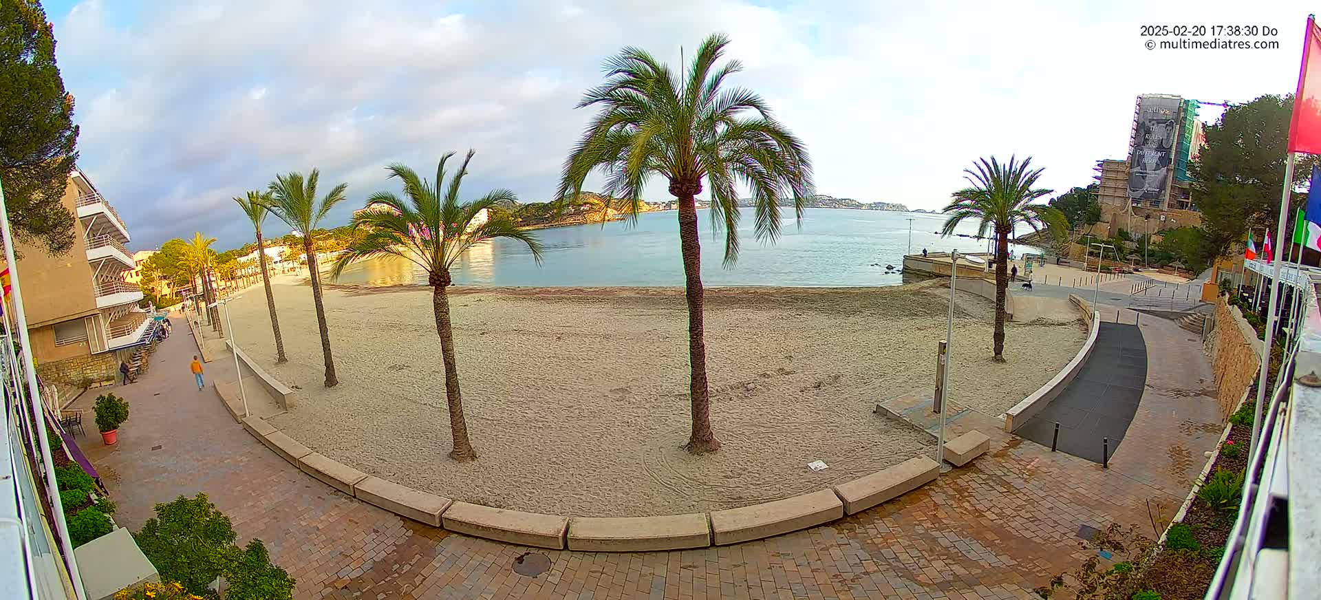 A mostly sunny day overlooks a sandy beach with palm trees, a paved walkway, and buildings in the background.