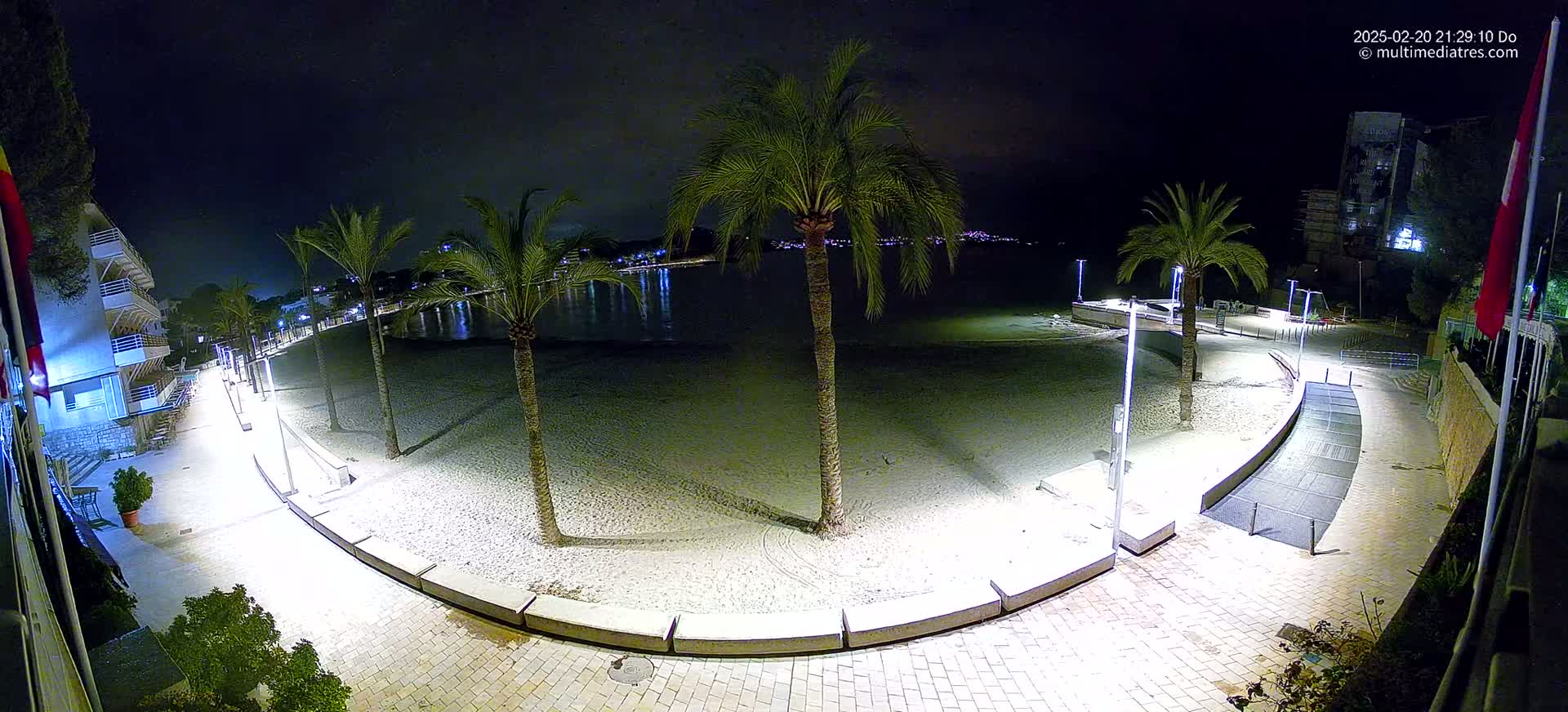 A nighttime view of a sandy beach bordered by a paved walkway lined with palm trees and lights, overlooking a calm body of water with city lights in the distance.