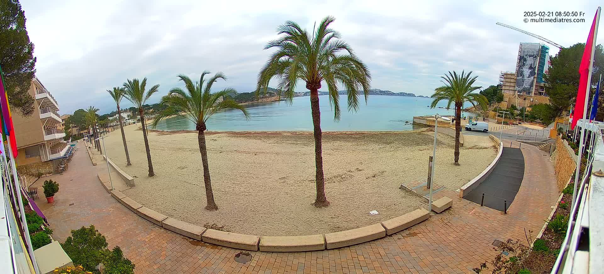A mostly empty sandy beach with palm trees, a paved walkway, and buildings is seen on an overcast day.