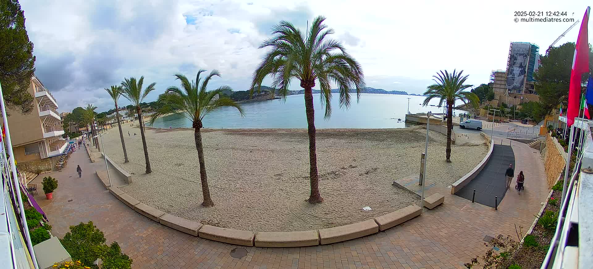 A sandy beach with palm trees is visible next to a paved walkway, overlooking calm water under a partly cloudy sky.