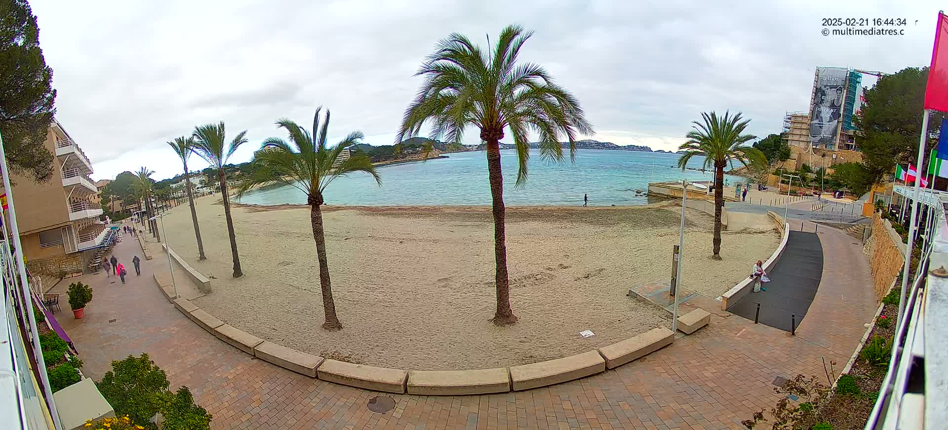 A sandy beach with palm trees and a paved walkway is visible, overlooking calm water under an overcast sky.