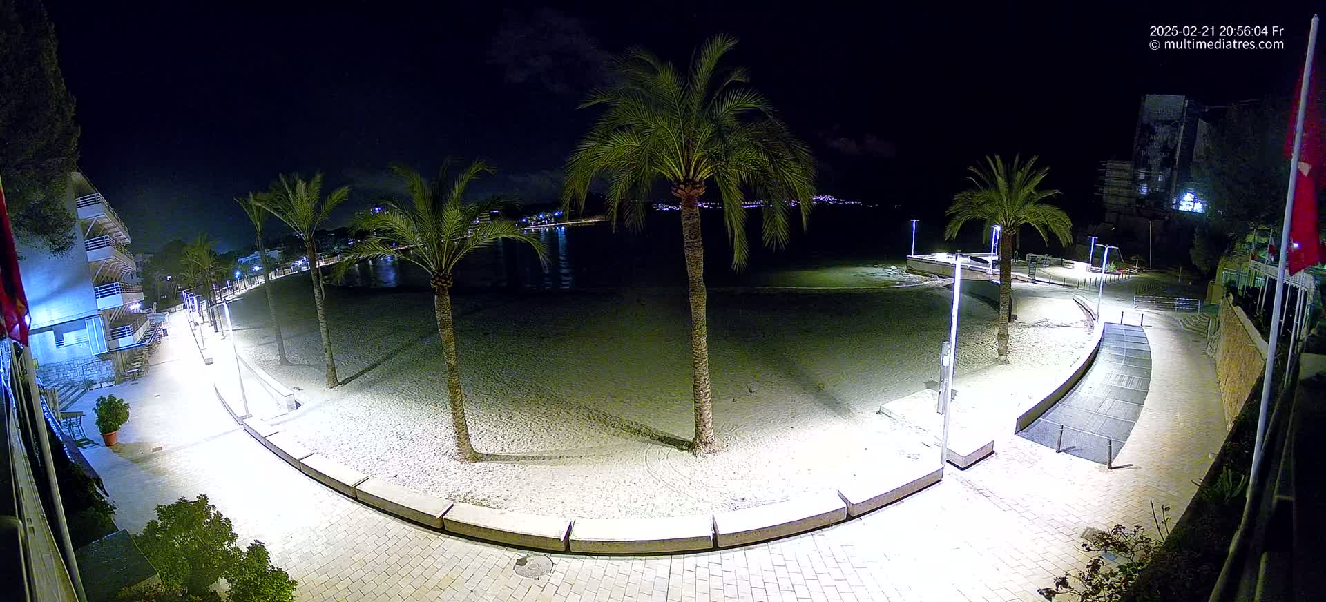 A nighttime view of a beachside promenade illuminated by lights, with palm trees lining the sandy beach and a calm body of water visible in the background.