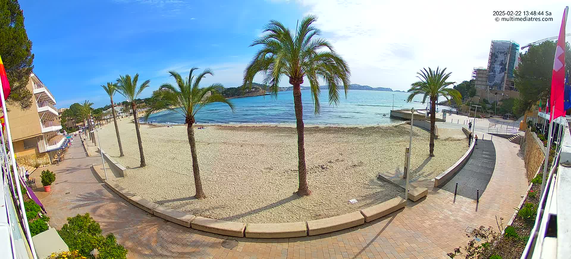 A sandy beach with palm trees and a paved walkway overlooks a calm, blue ocean under a sunny sky.