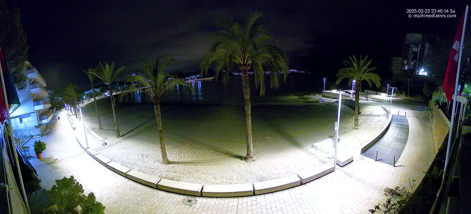 A nighttime, brightly lit beach scene shows a sandy beach, palm trees, a paved walkway, and buildings in the background.