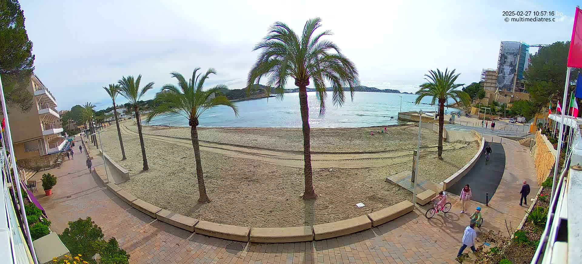 A sandy beach with palm trees lines a paved walkway, overlooking calm ocean water under a mostly clear sky, with several people walking and cycling along the path.