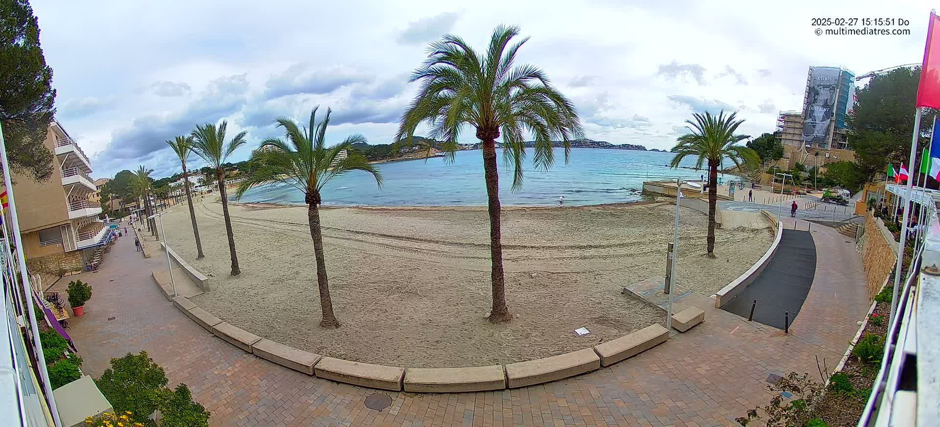 A sandy beach with palm trees lines a paved walkway next to a calm, blue sea under a partly cloudy sky.