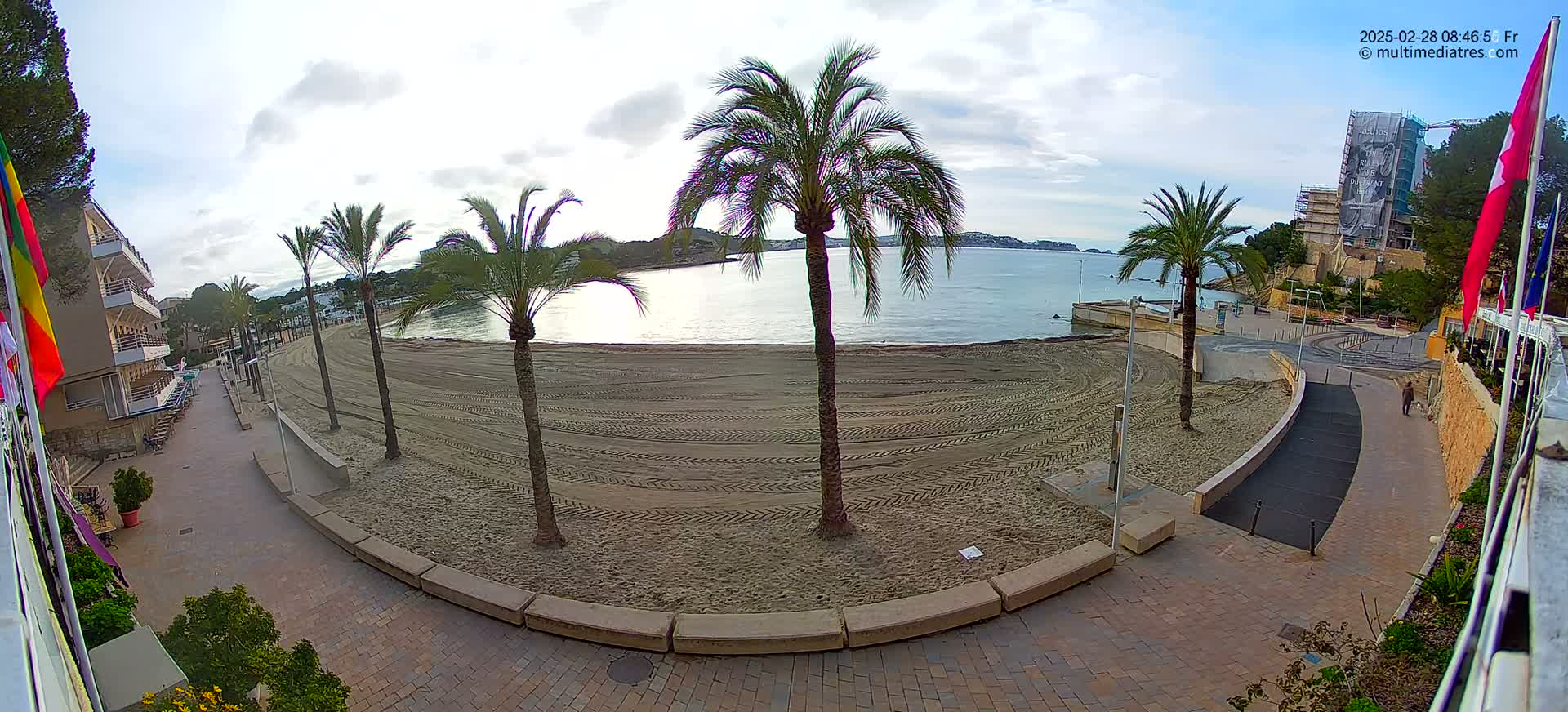 A mostly empty sandy beach with palm trees is viewed from a high vantage point on a partly cloudy day, with a walkway and buildings visible in the background.