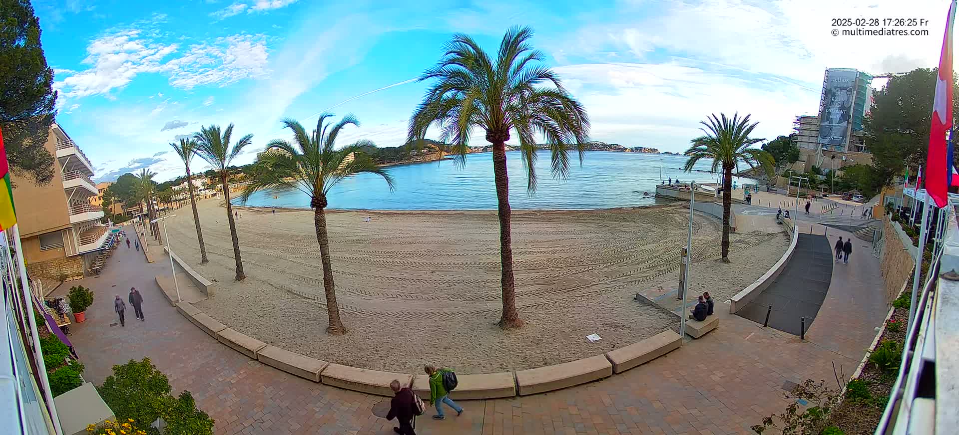 A mostly sunny day at a sandy beach, featuring palm trees, a paved walkway, and a few people strolling along the shore.