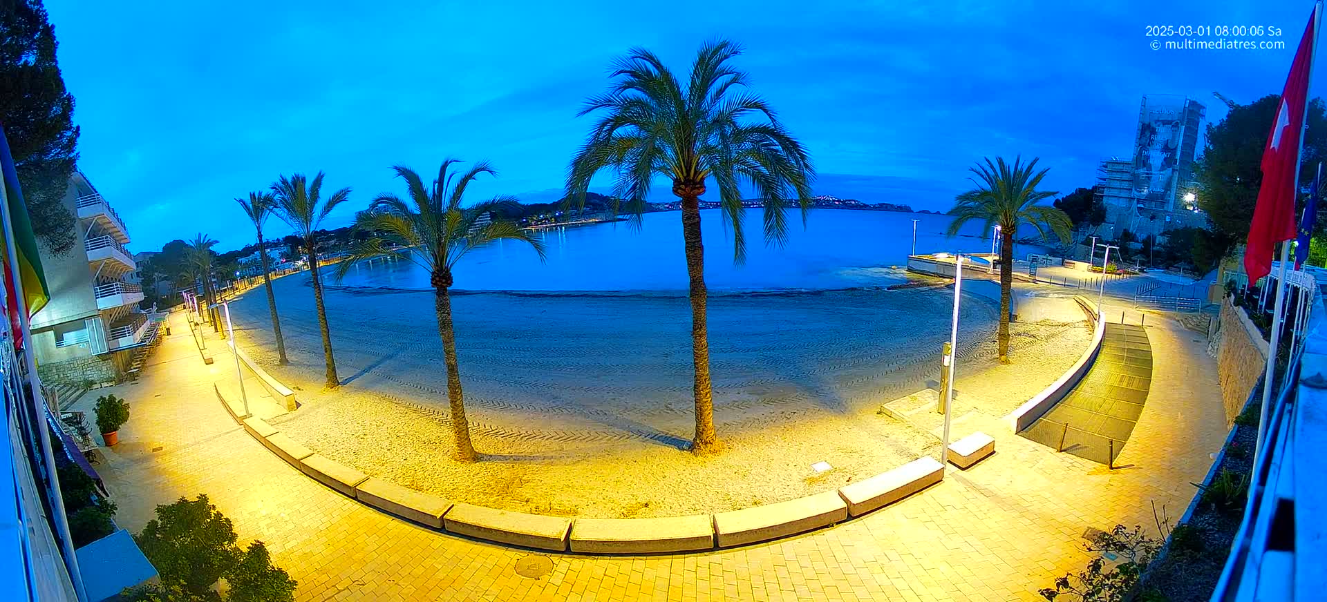 A mostly empty sandy beach at twilight is illuminated by artificial lights, with palm trees lining a paved walkway leading to a calm body of water.