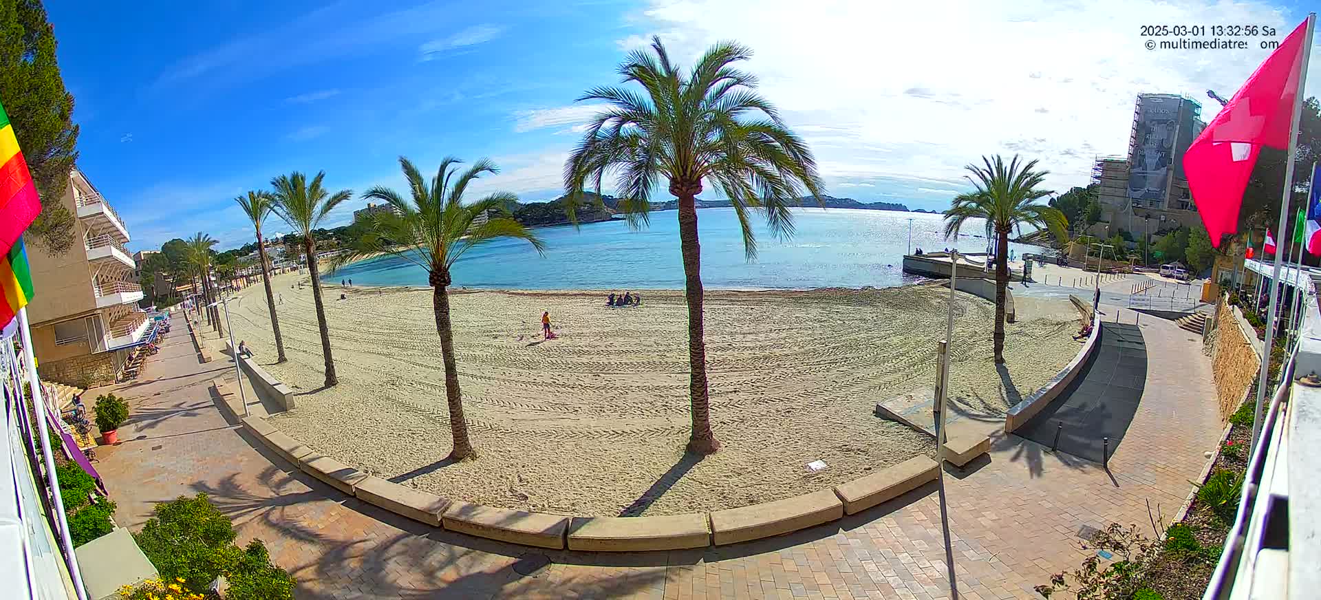 A sandy beach with palm trees and a paved walkway is seen on a sunny day, overlooking a calm body of water.