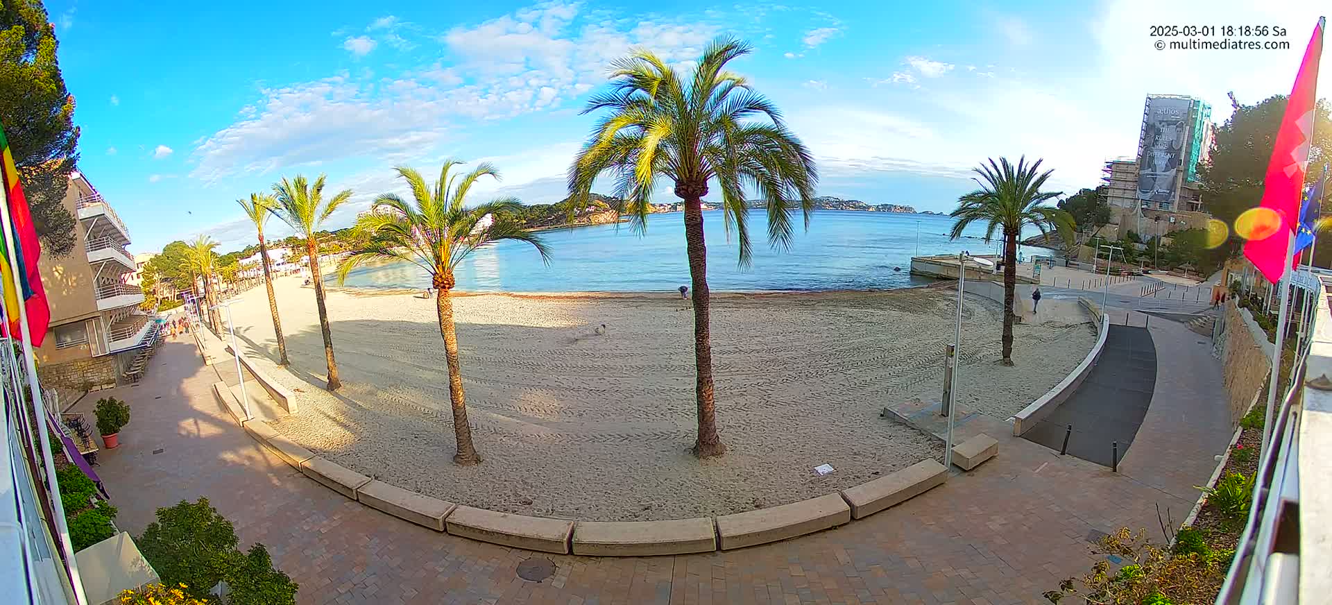 A mostly empty sandy beach with palm trees and a paved walkway, overlooking calm blue water under a partly cloudy sky.