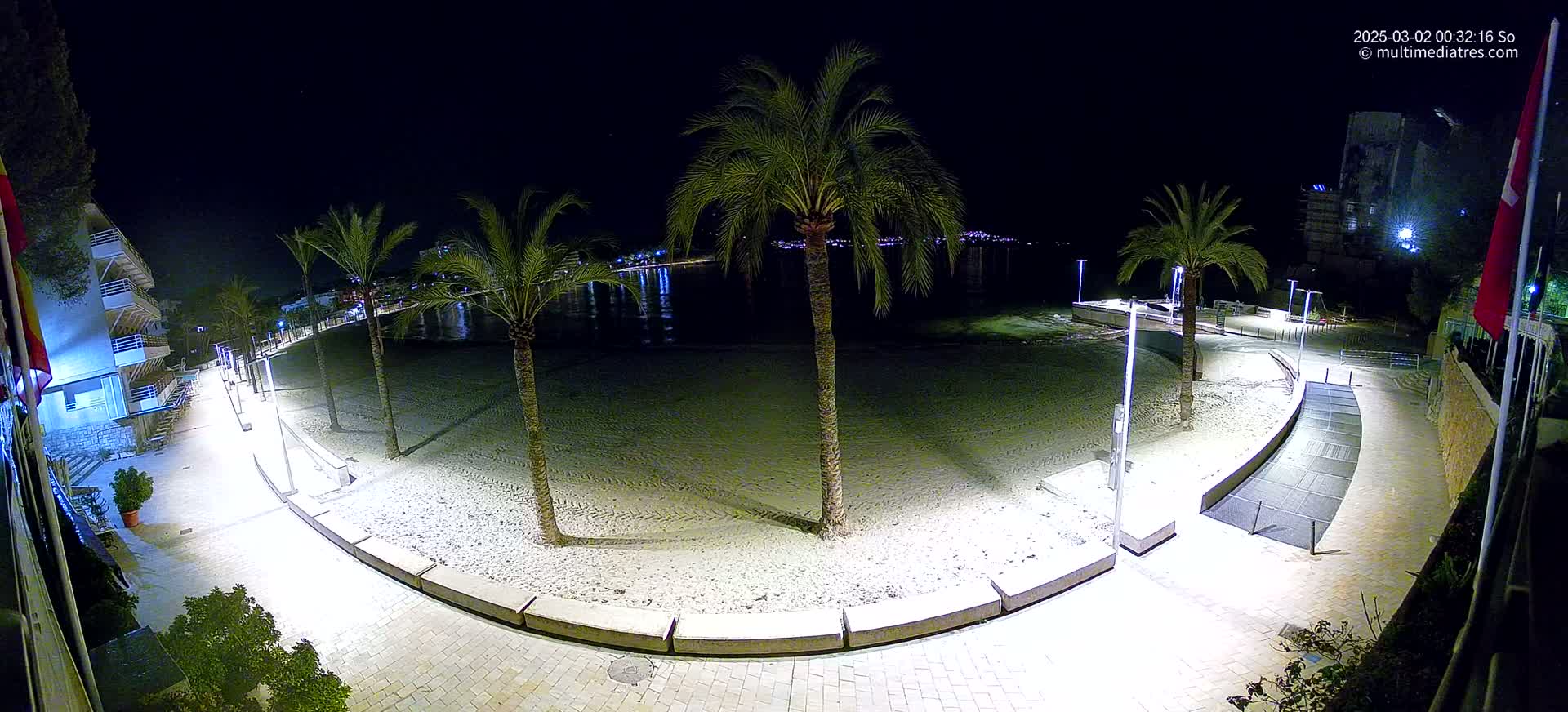 A nighttime view of a sandy beach with palm trees, illuminated walkways, and buildings in the background.