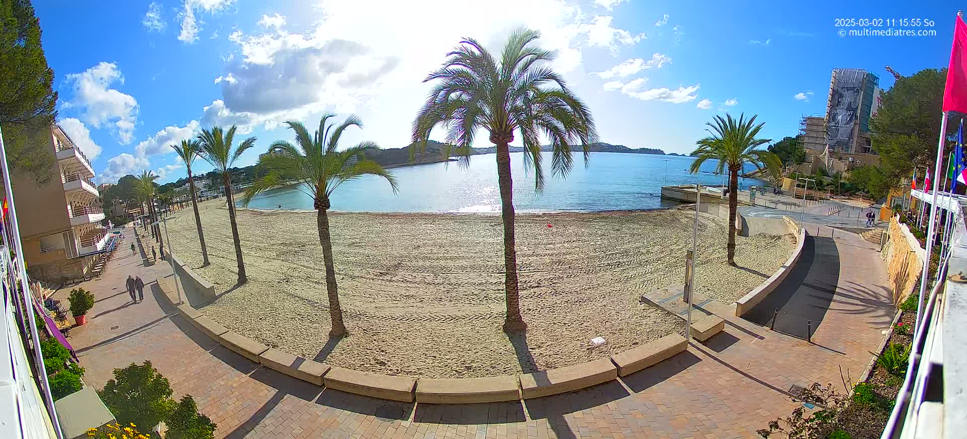 A sandy beach with palm trees and a paved walkway is seen on a sunny day with partly cloudy skies, overlooking calm blue water.