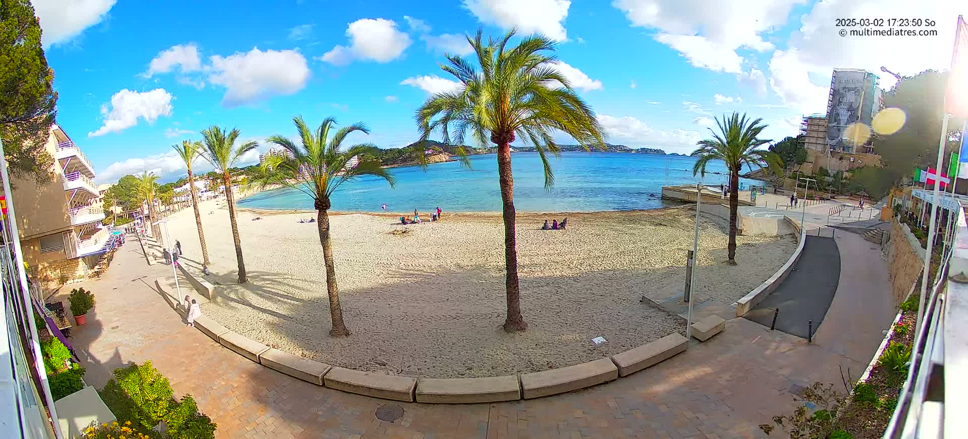 A sandy beach with palm trees and a few people is situated next to a paved walkway and buildings, under a mostly sunny sky with some scattered clouds.