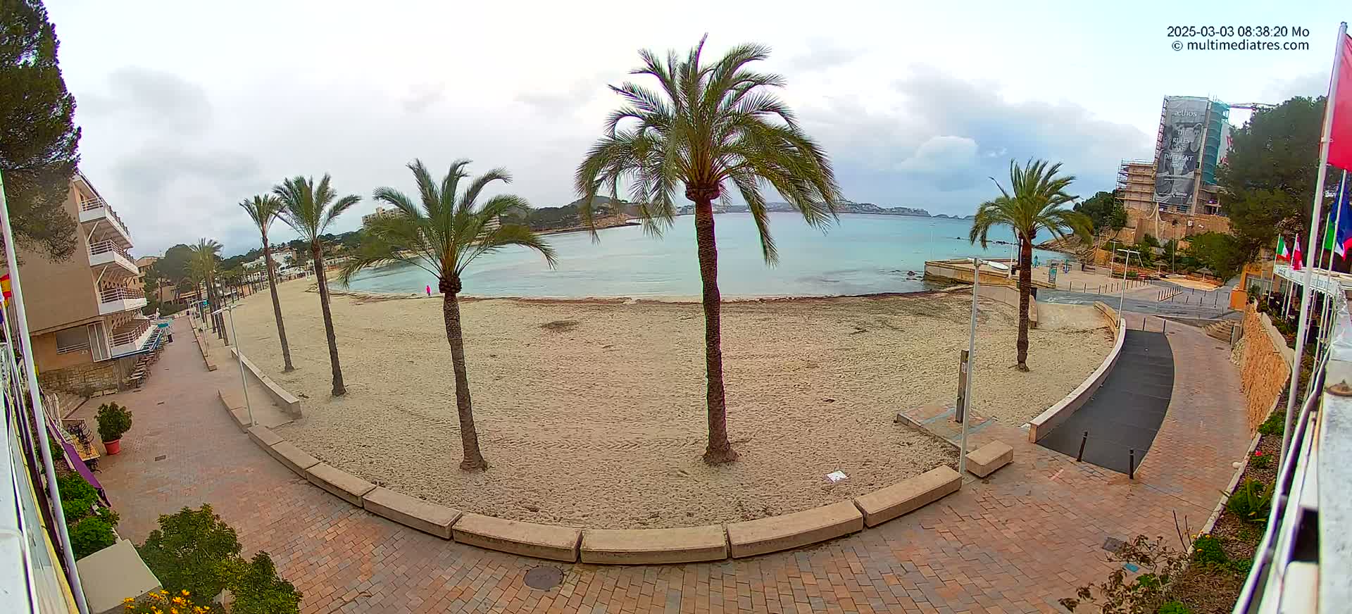 A sandy beach with palm trees and a brick walkway is visible, under an overcast sky, next to a calm body of water.